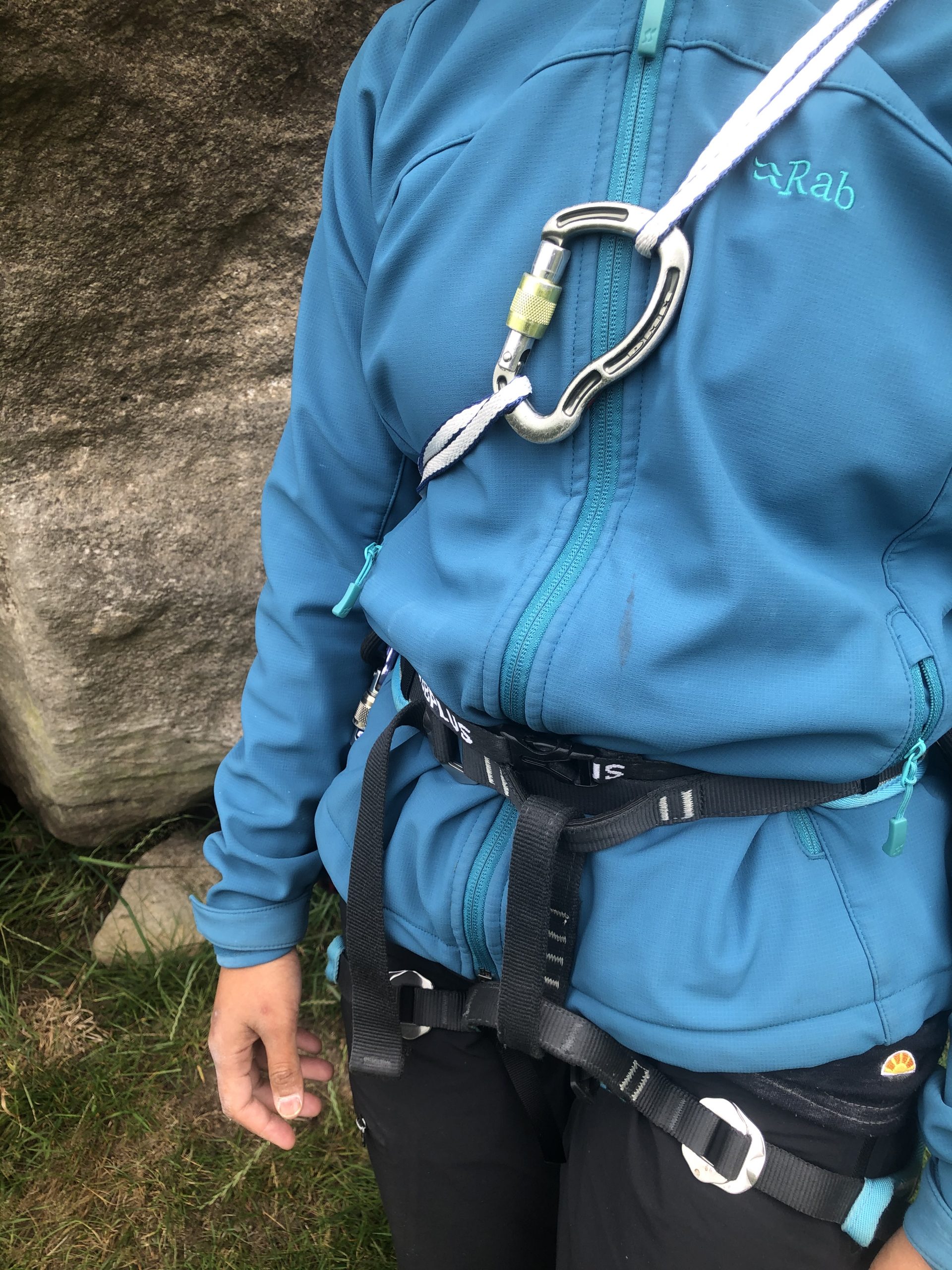 A close up showing an effective way of carrying a climbing sling while climbing - it is run around the neck and underneath one armpit and clipped together with a karabiner ” width=