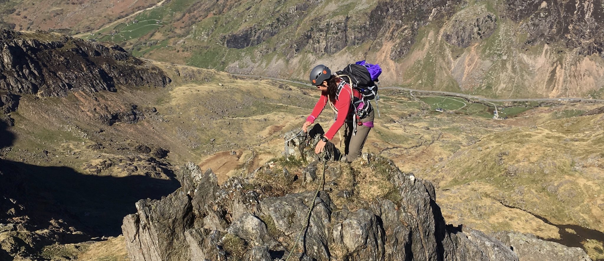 A mountaineer reaches the crest of the ridge of the Clogwyn y Person Arete with Llanberis Pass below during an advanced scrambling course in Snowdonia” width=