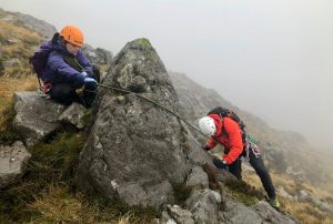 One mountaineer uses the rope around a huge rocks a direct belay to safeguard her partner during a learn to lead scrambling course in Snowdonia