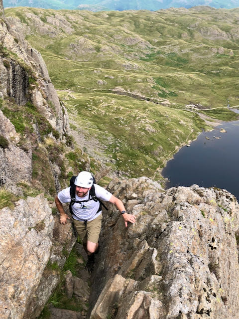 A scrambler climbing high on Jack's Rake, a Grade 1 scramble in the Lake District, with Stickle Tarn far below him